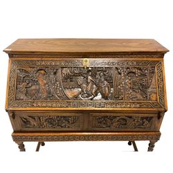 Mid-to late 20th century Singapore camphor wood bureau on stand, the fall front carved with royal procession scene within a cityscape, flower head carved border, the interior fitted with drawer and correspondence shelves, two drawers below, on turned supports united by H-shaped stretchers