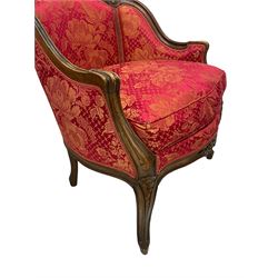 Late 20th century French design stained beech framed armchair and matching stool - the armchair with shaped and moulded frame carved with flower heads and foliage, upholstered in silk red and gold ground fabric with floral pattern, on scroll carved cabriole feet decorated with flower head carvings (W73cm, H82cm, D75cm); the footstool of concaved rectangular form with moulded frame carved with flower heads, upholstered loose cushion, on cabriole feet (70cm x 64cm, H48cm)