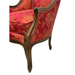 Late 20th century French design stained beech framed armchair and matching stool - the armchair with shaped and moulded frame carved with flower heads and foliage, upholstered in silk red and gold ground fabric with floral pattern, on scroll carved cabriole feet decorated with flower head carvings (W73cm, H82cm, D75cm); the footstool of concaved rectangular form with moulded frame carved with flower heads, upholstered loose cushion, on cabriole feet (70cm x 64cm, H48cm)